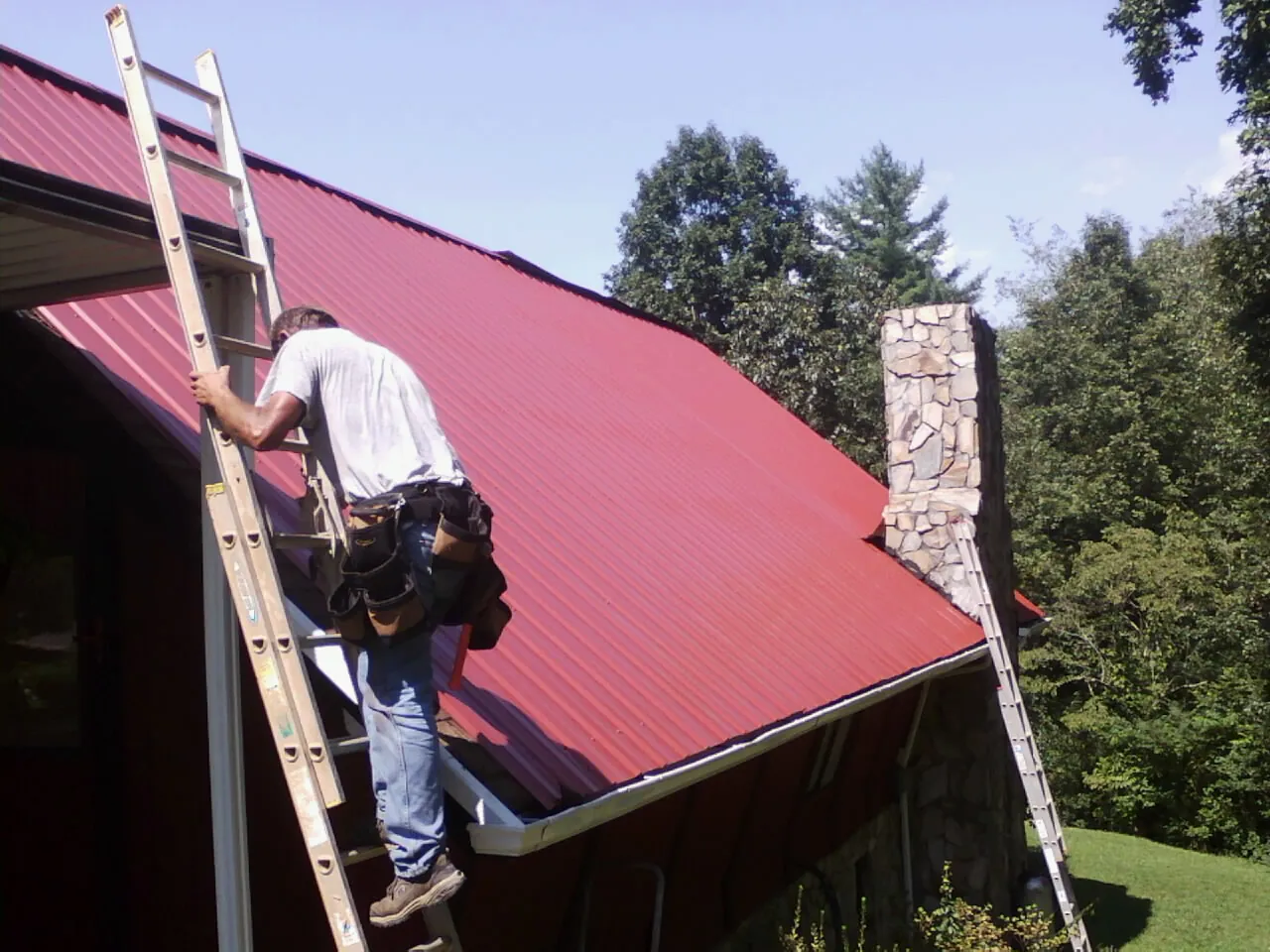 Roofer on a ladder beside a red metal roof and stone chimney — Soffit & Fascia Repair by Roofing Brothers in Medaryville